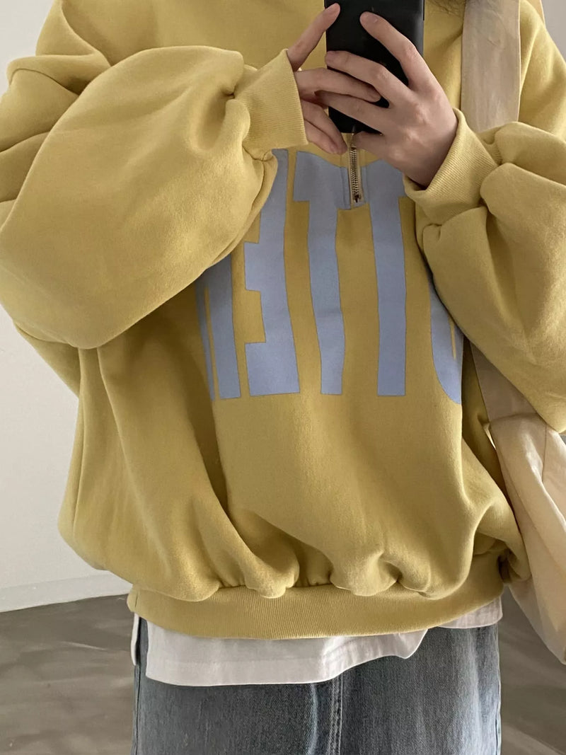 A person wearing an oversized mustard yellow sweatshirt with a light blue graphic print on the front, styled over a white t-shirt and blue jeans.