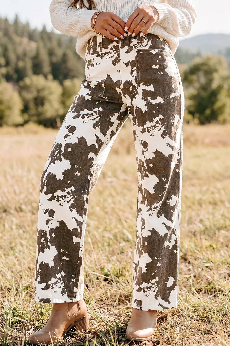 A person standing outdoors in a grassy field wearing wide-leg corduroy pants with a brown and off-white mottled pattern, paired with a cream sweater and tan heeled boots.