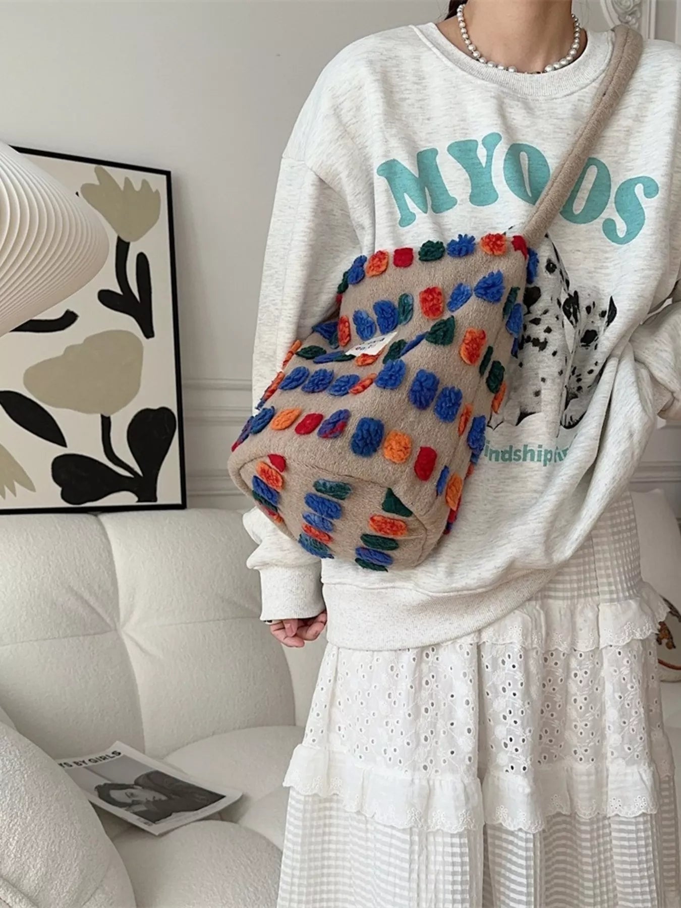 Person wearing a white sweatshirt with colorful text and holding a multicolored textured bag in a room.