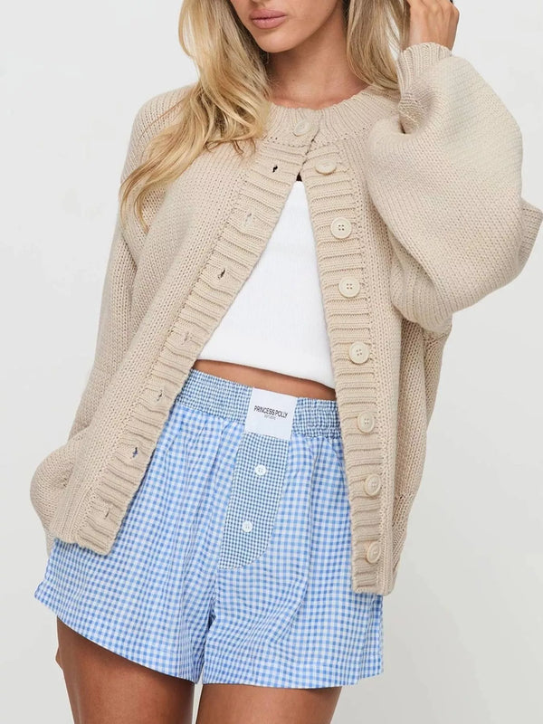 A woman wearing a beige chunky knit cardigan with large buttons over a white crop top and blue-and-white gingham shorts, photographed against a plain light background.