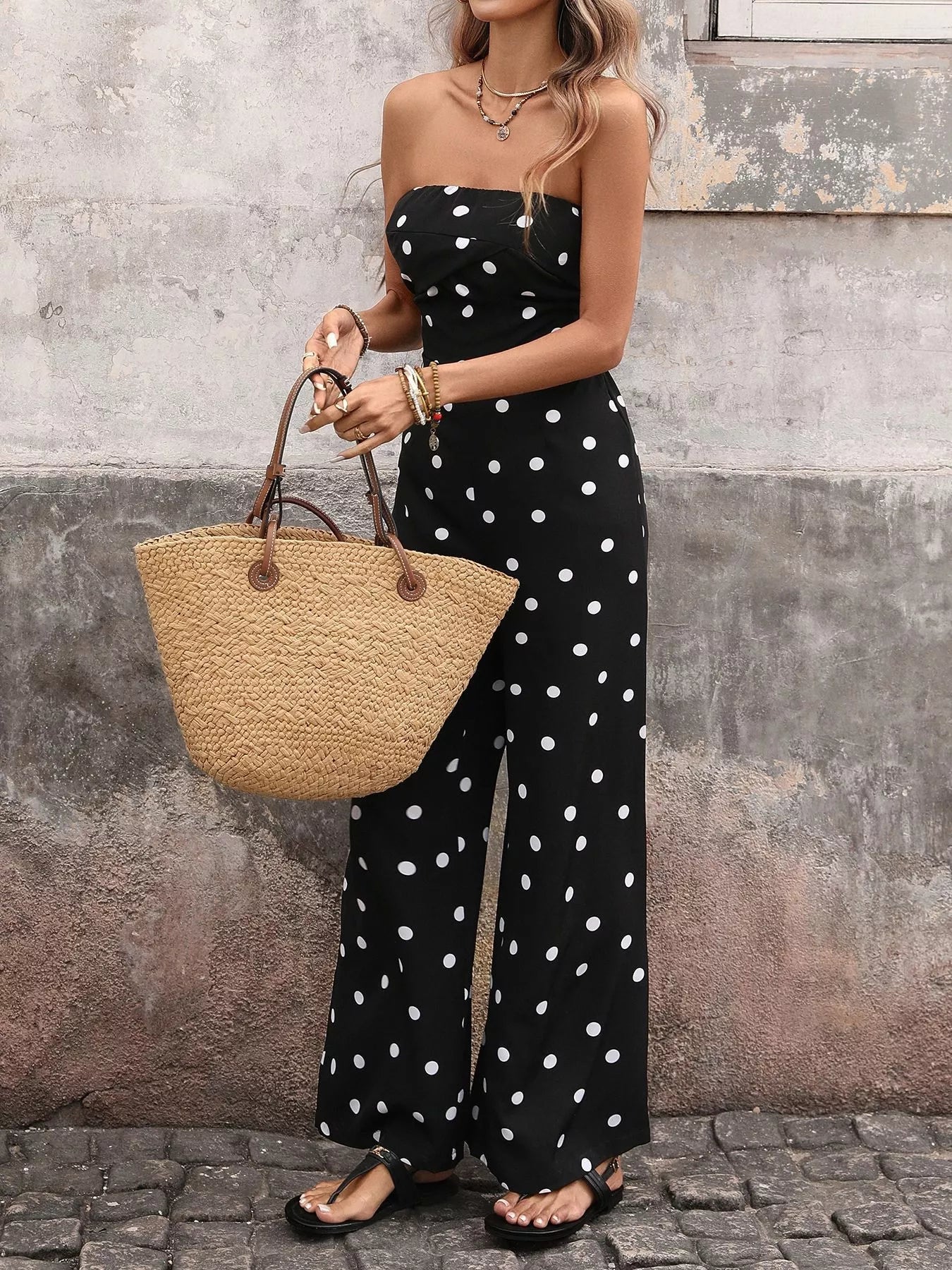 Woman wearing a black polka dot jumpsuit holding a straw bag against a textured wall.