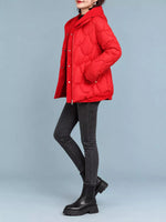 A woman wearing a bright red quilted puffer jacket with a high collar, paired with black skinny jeans and black ankle boots, standing against a solid light gray studio background.