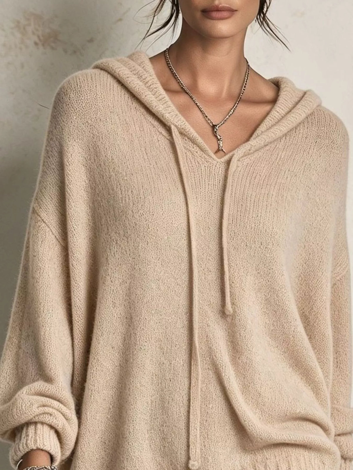A close-up of a model wearing a soft, oversized knit hoodie in a warm beige or cream color. The hoodie features a V-neckline, drawstring hood, and ribbed cuffs and hem.