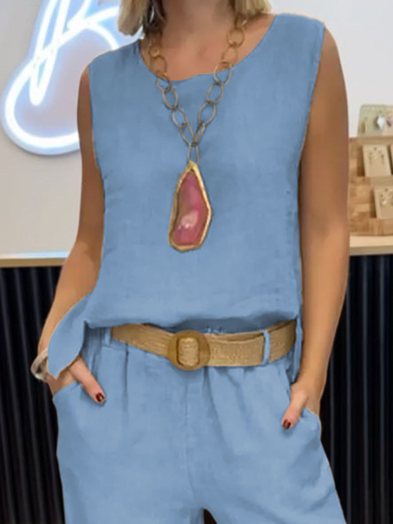 Person wearing a blue sleeveless top and pants with a large necklace, standing indoors.