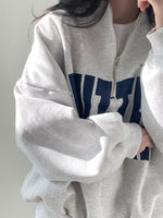 A person wearing an oversized light gray quarter-zip sweatshirt with a large navy blue block graphic across the chest, layered over a white t-shirt.