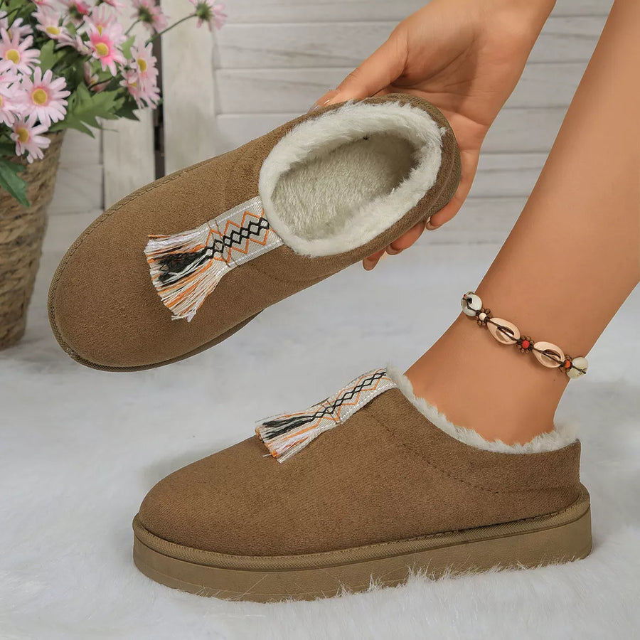 Brown slippers with tassel details held by a hand on a light background