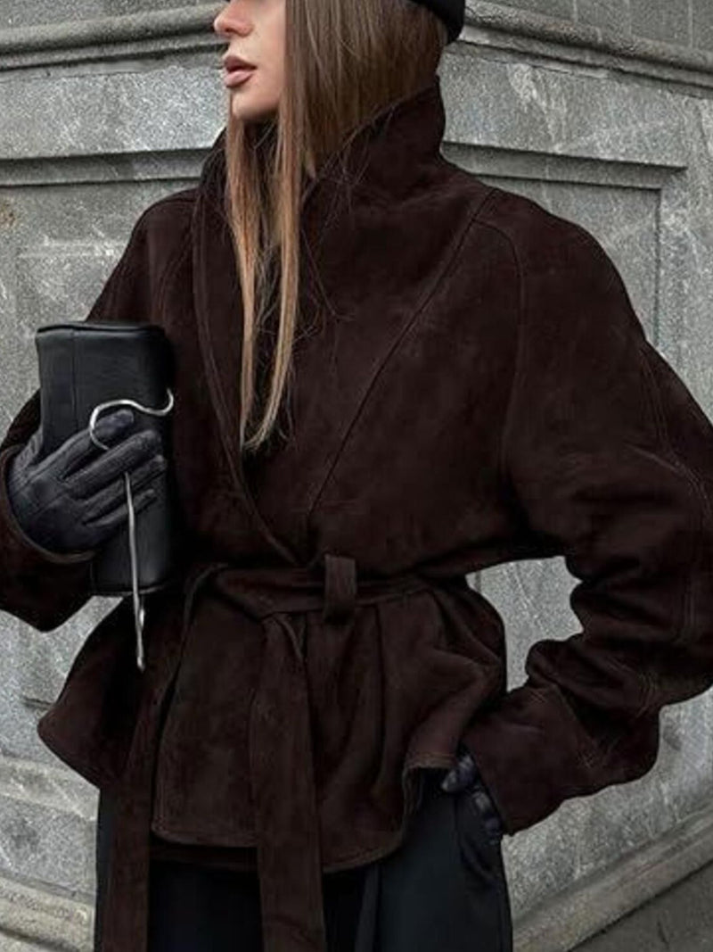 Person wearing a brown coat with a belt, holding a black bag against a gray wall.