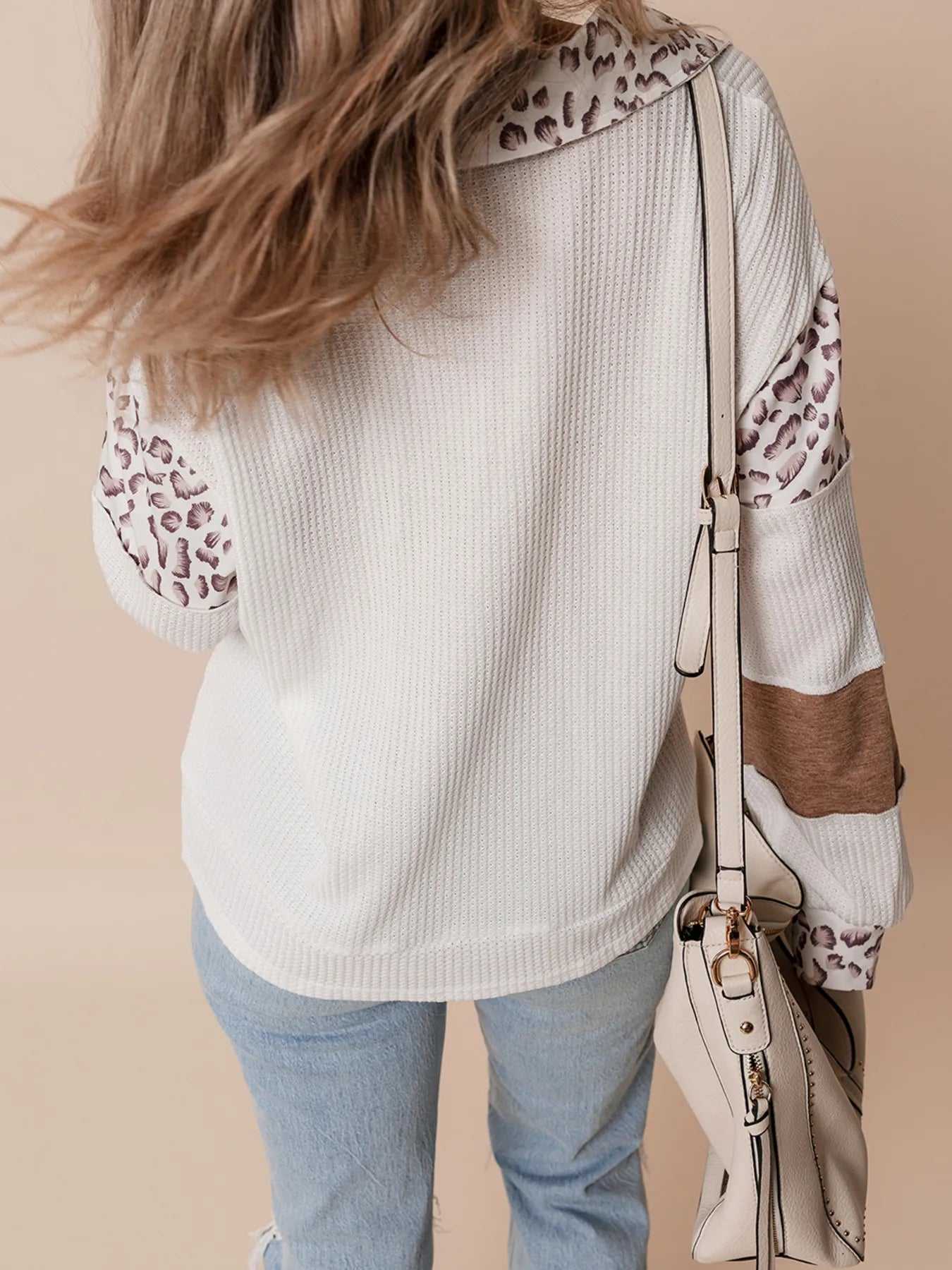 Person wearing a white sweater with leopard print sleeves and holding a beige handbag against a beige background
