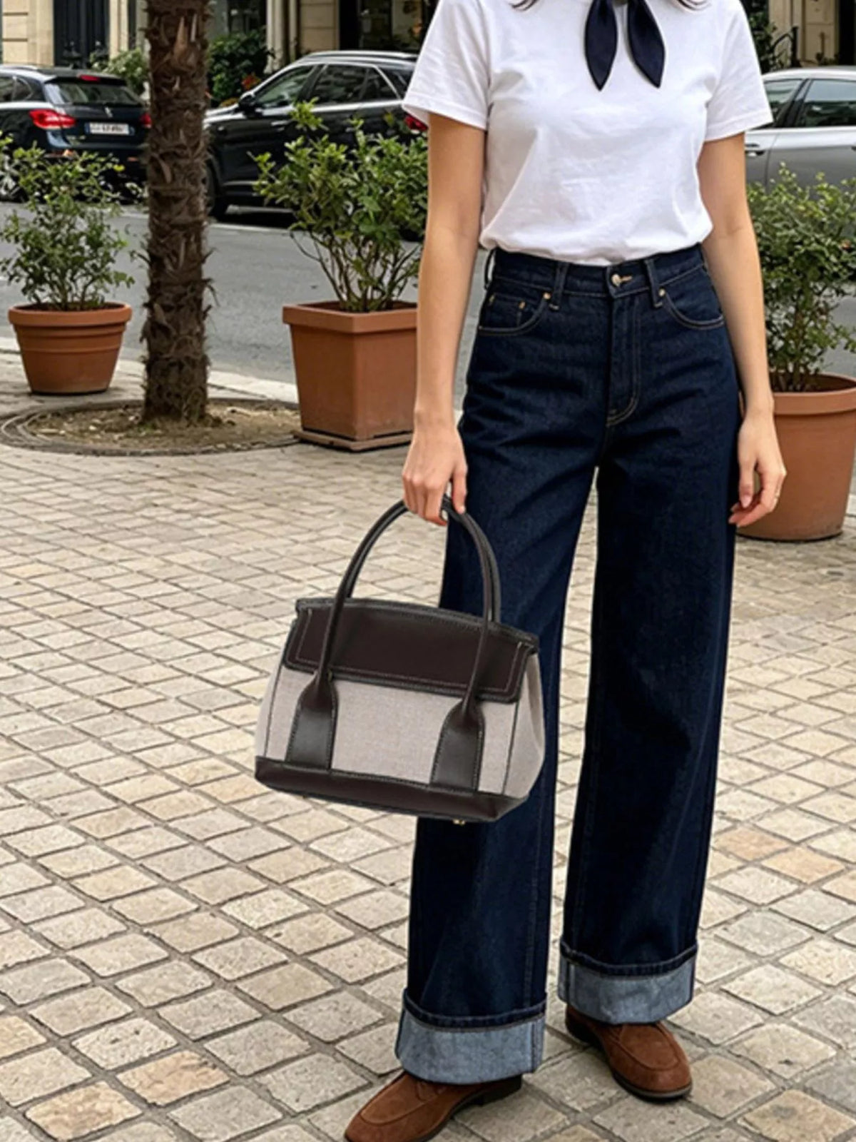 A person standing outdoors on a paved street holding a structured handbag. The person is wearing a white short-sleeve top with a dark neck scarf, dark blue wide-leg jeans with rolled cuffs, and brown shoes.