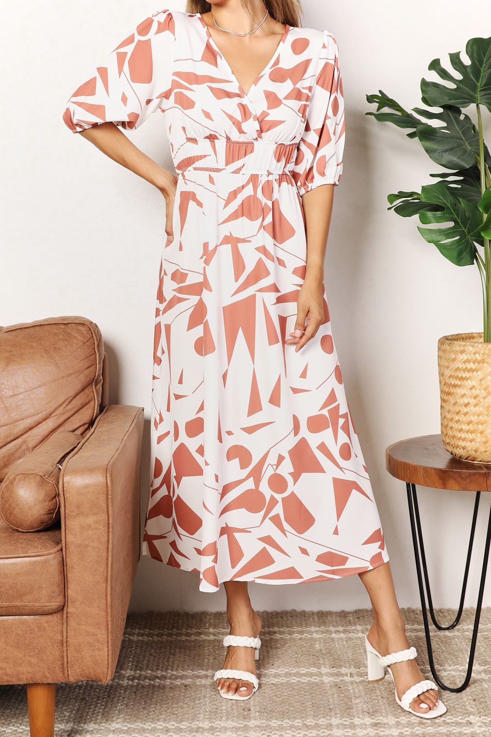 Woman wearing a patterned dress standing in a room with a brown leather chair and a plant.