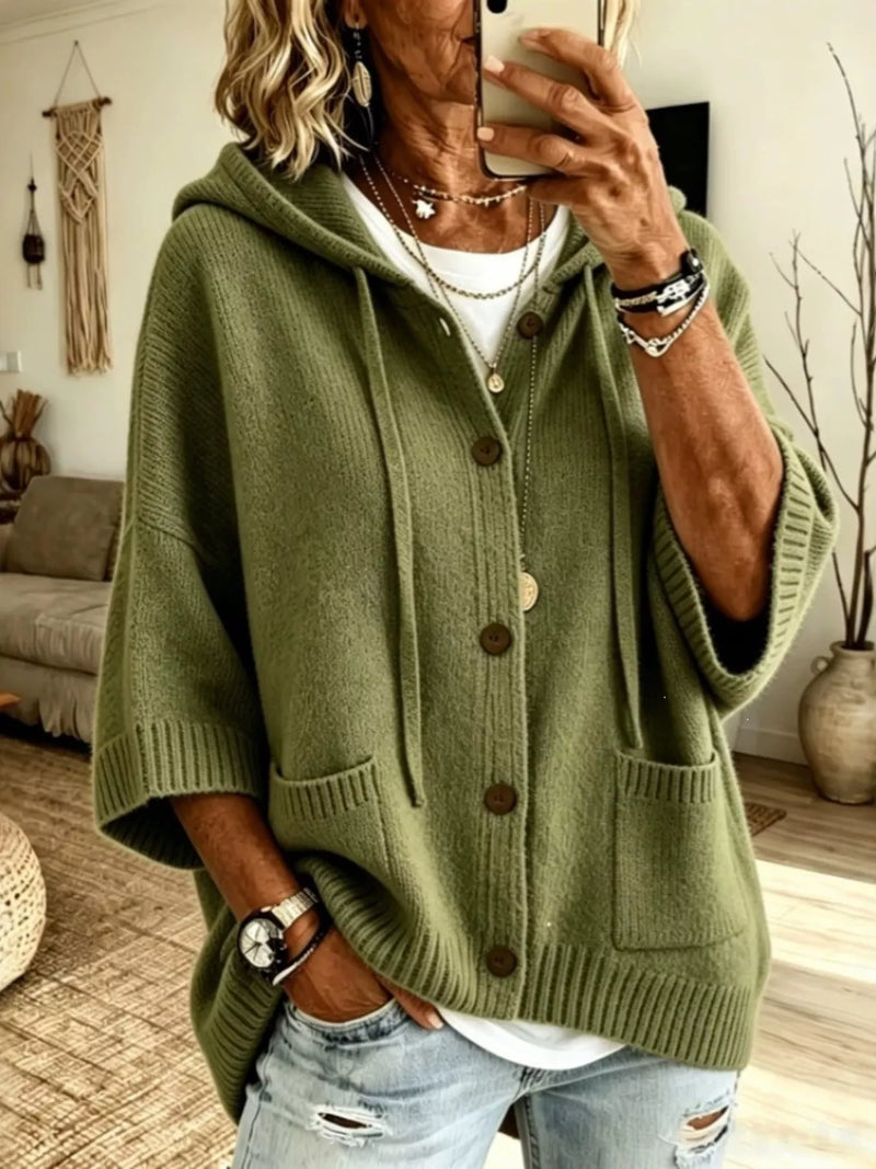 A woman wearing an olive green knit hooded cardigan with front buttons and large patch pockets, paired with light blue distressed jeans, taking a mirror selfie in a living room setting.