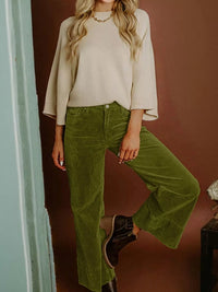 Person wearing a beige top and green pants against a brown wall.