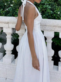 Woman wearing a white dress with a halter neck design, standing against a stone railing with greenery in the background.