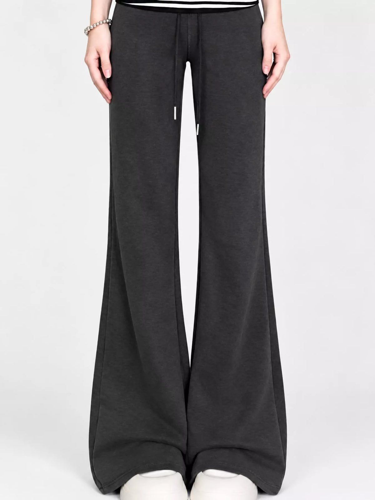 A person wearing dark charcoal wide-leg sweatpants with a drawstring waist, paired with a black and white top and white sneakers, shown against a light gray studio background.
