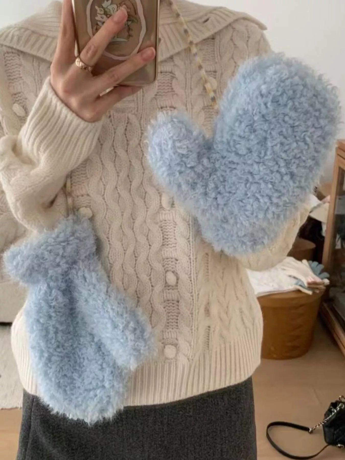 Person wearing a beige cable knit sweater and holding blue fuzzy gloves.