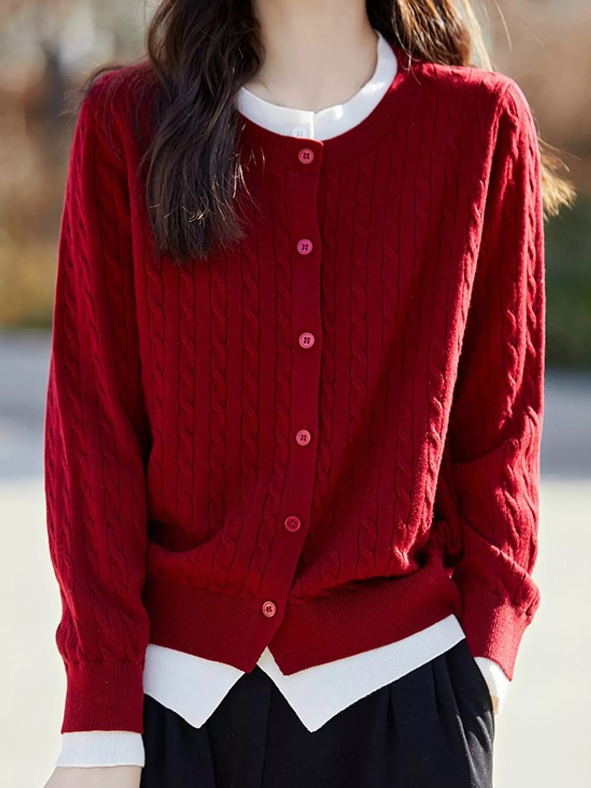 A woman wearing a red cable-knit button-up cardigan over a white collared shirt and black skirt, photographed outdoors with a blurred background.