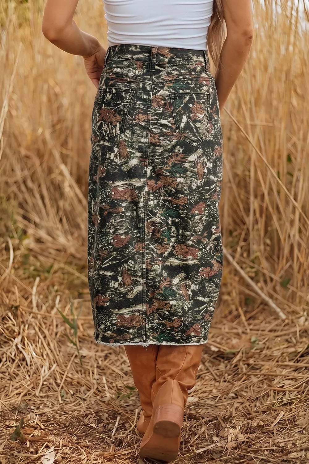 A person outdoors in a field of dry grass, wearing a camouflage midi skirt with a white top and brown boots.