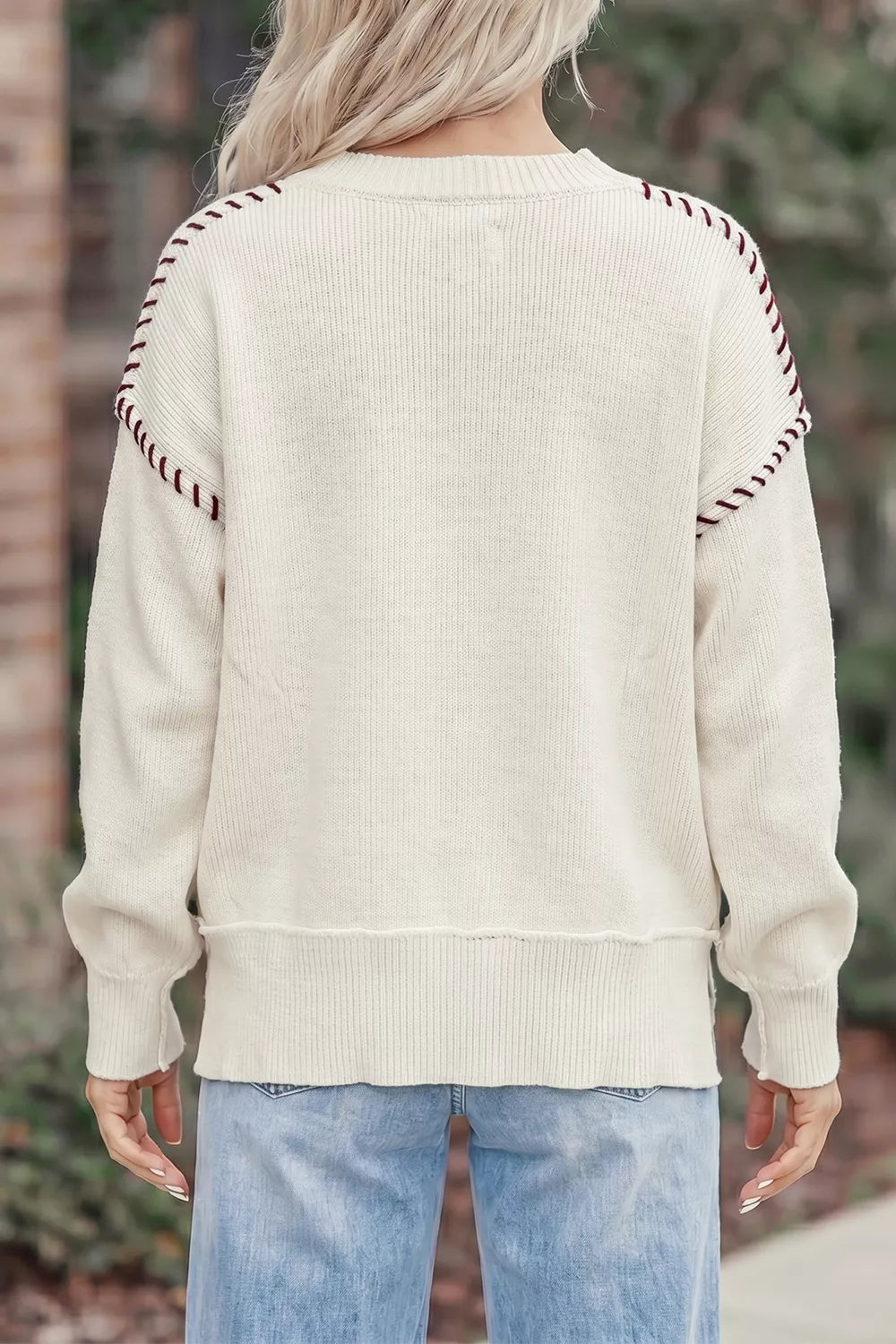 A person viewed from the back wearing a cream ribbed knit sweater with decorative dark red and white striped trim along the shoulders, paired with light blue jeans.
