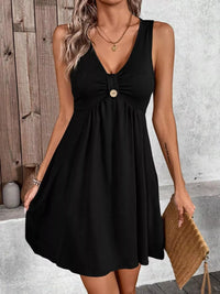 Woman wearing a black sleeveless dress with a button detail, holding a beige clutch against a neutral background.