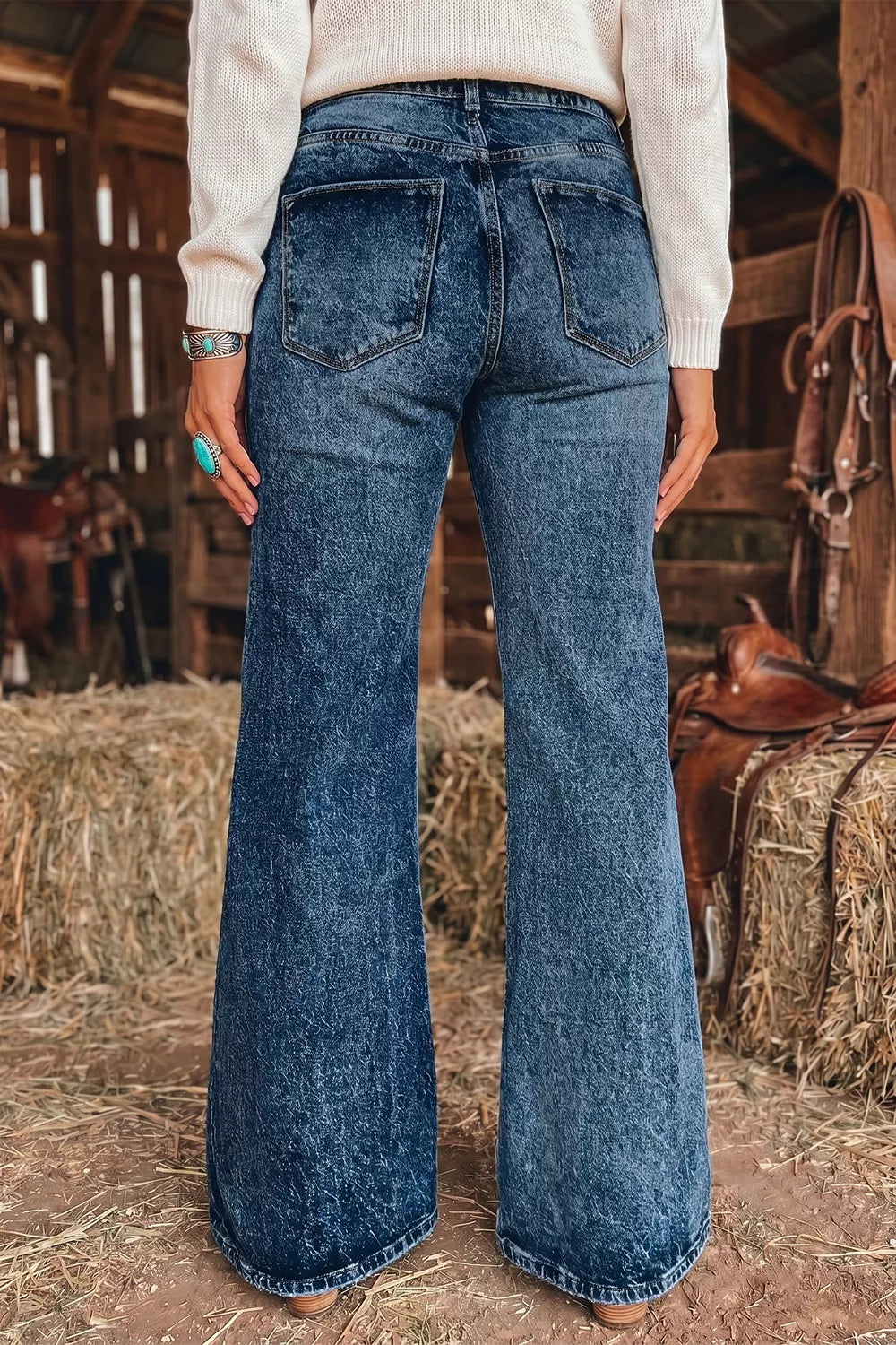 A person viewed from the back wearing dark blue wide-leg denim jeans in a barn setting with hay bales and tack visible in the background.