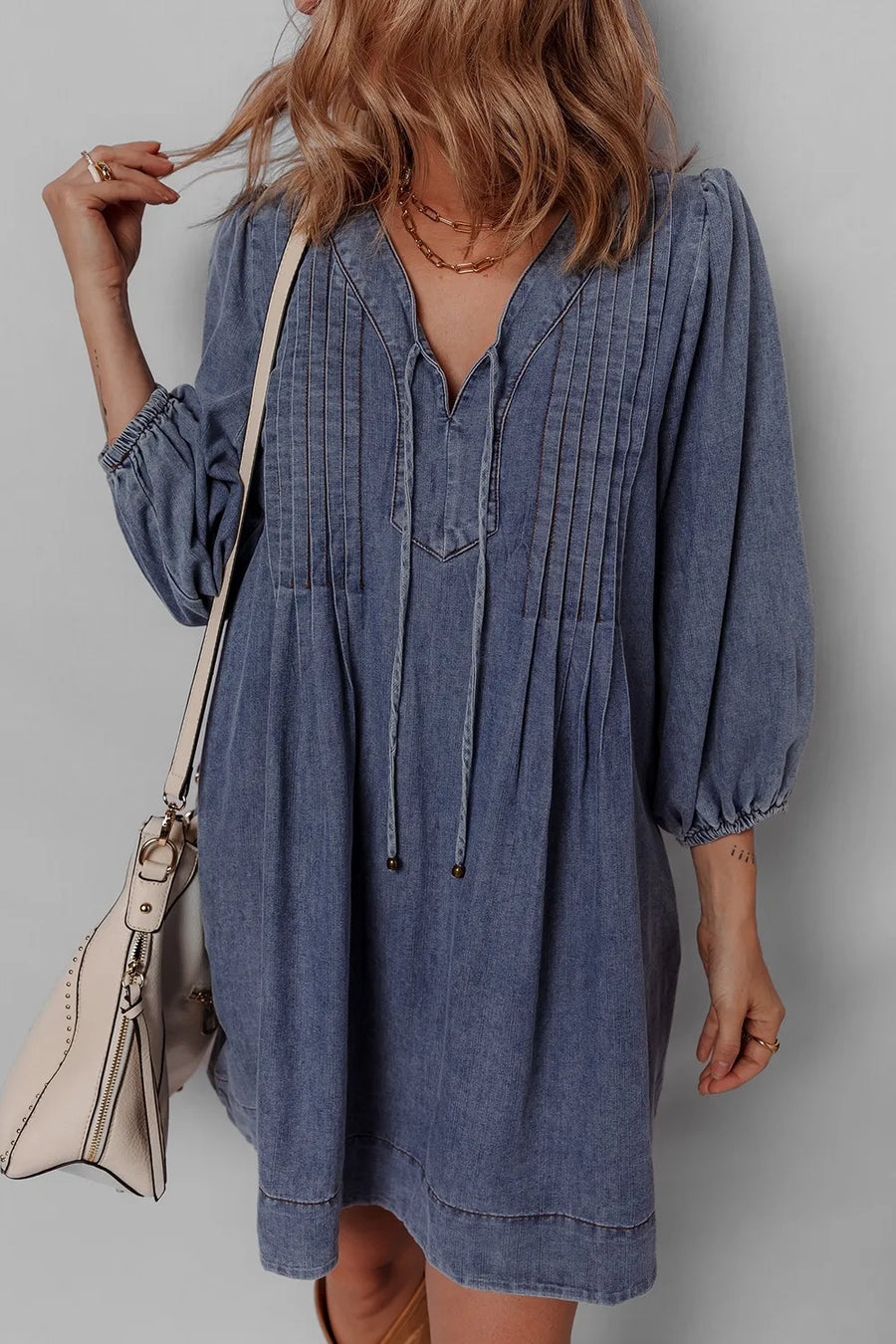 Woman wearing a blue denim dress with a beige handbag against a plain background
