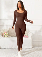 Woman wearing a brown bodysuit in a white room with a couch and flowers.