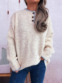 Person wearing a beige sweater with button details and blue jeans against a light purple wall.