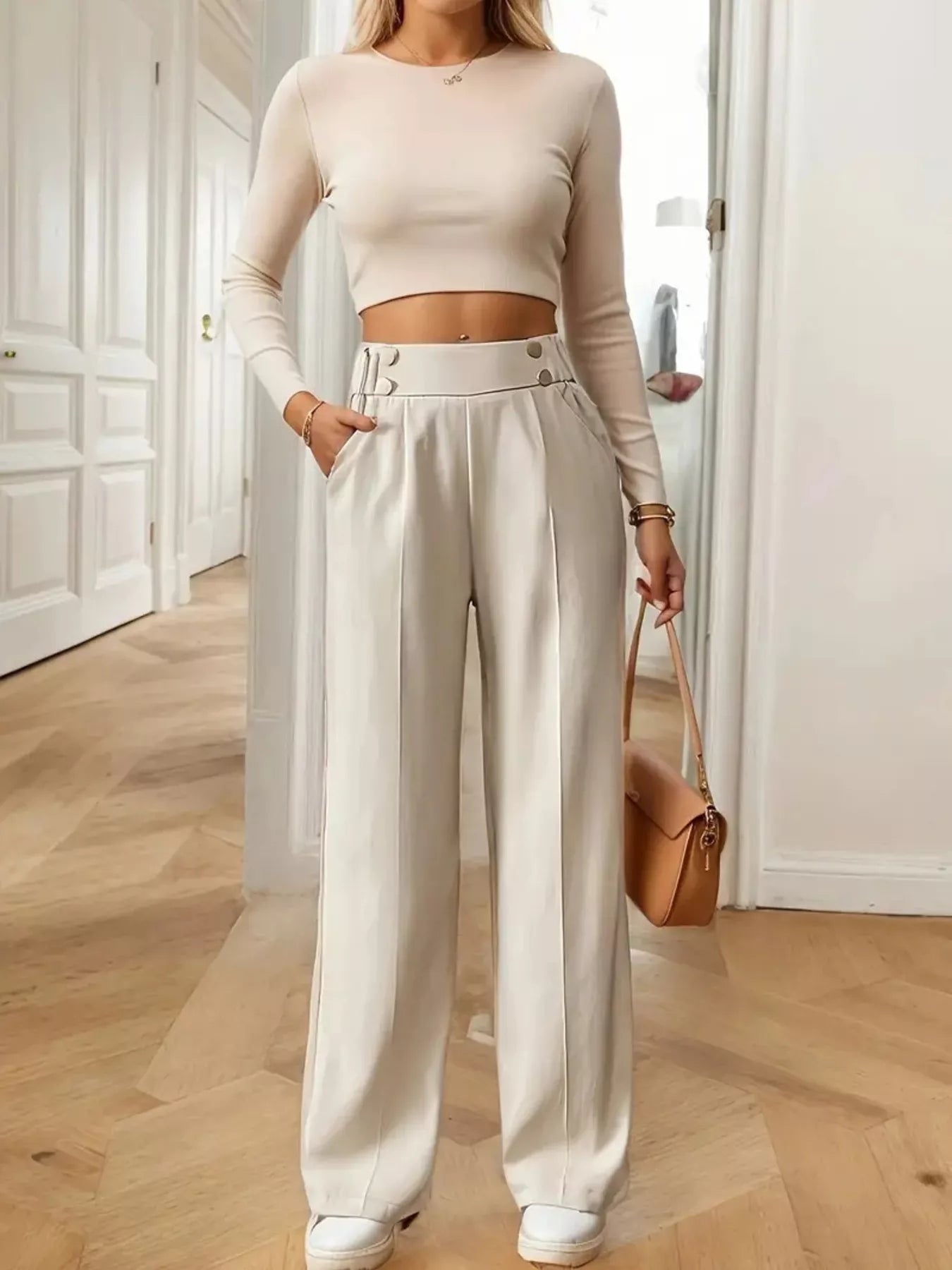 Person wearing a beige long-sleeve top and white wide-leg pants, holding a brown handbag indoors.