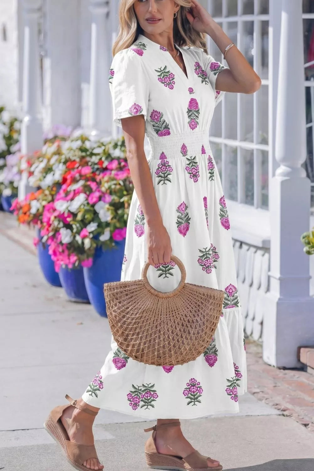 Woman in a white dress with floral patterns holding a straw bag, standing on a sidewalk with flowers in the background.