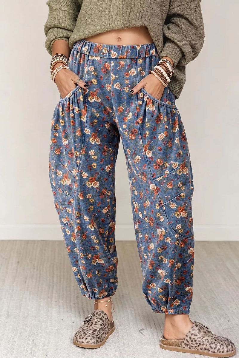 A person wearing loose, elastic-waist floral pants in blue with orange, cream, and brown floral print, styled with an olive green cropped sweater and leopard-print slip-on shoes.