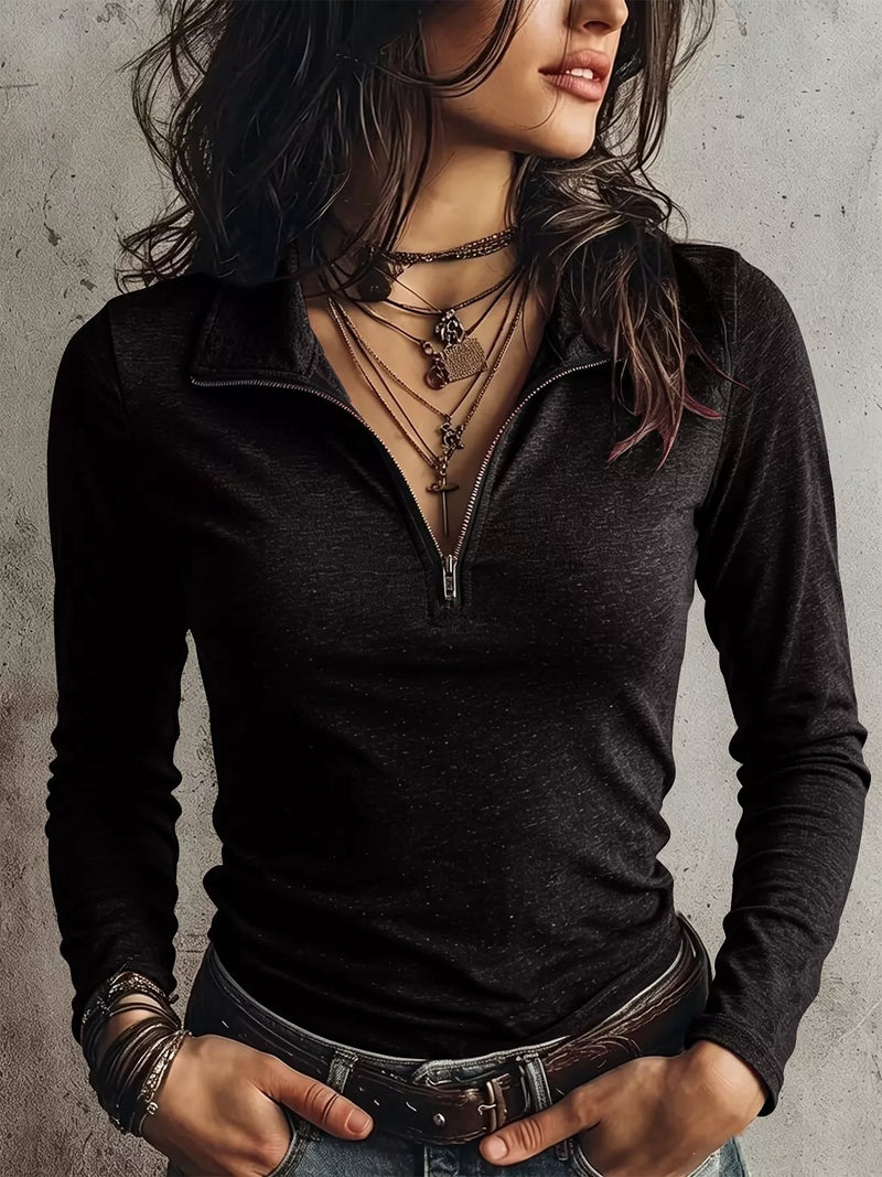 A woman wearing a fitted black long-sleeve quarter-zip polo shirt, styled with layered necklaces, bracelets, and a brown belt, standing against a textured gray concrete wall.