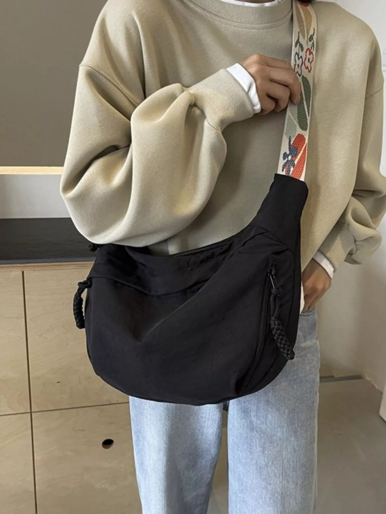 Person wearing a beige sweater and light blue jeans holding a black bag with a colorful strap.