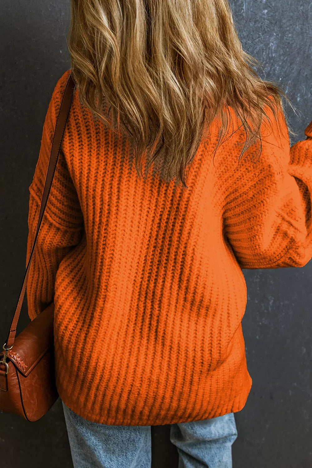 A person viewed from behind wearing a bright orange ribbed knit sweater and light blue jeans, with a brown shoulder bag, against a dark textured wall.