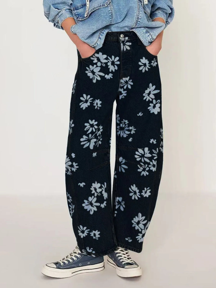 Person wearing black floral-patterned pants with a denim jacket on a plain background