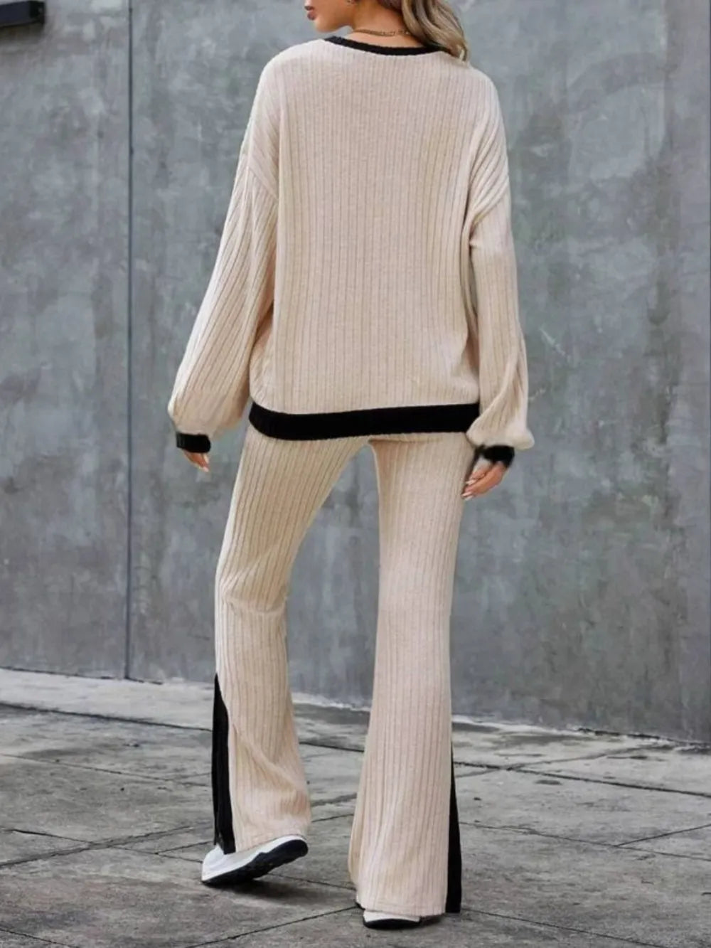Person wearing a beige ribbed sweater and pants with black trim against a gray wall.