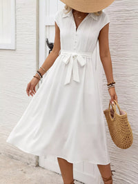 Woman wearing a white dress with a belt, holding a straw bag, against a light background.