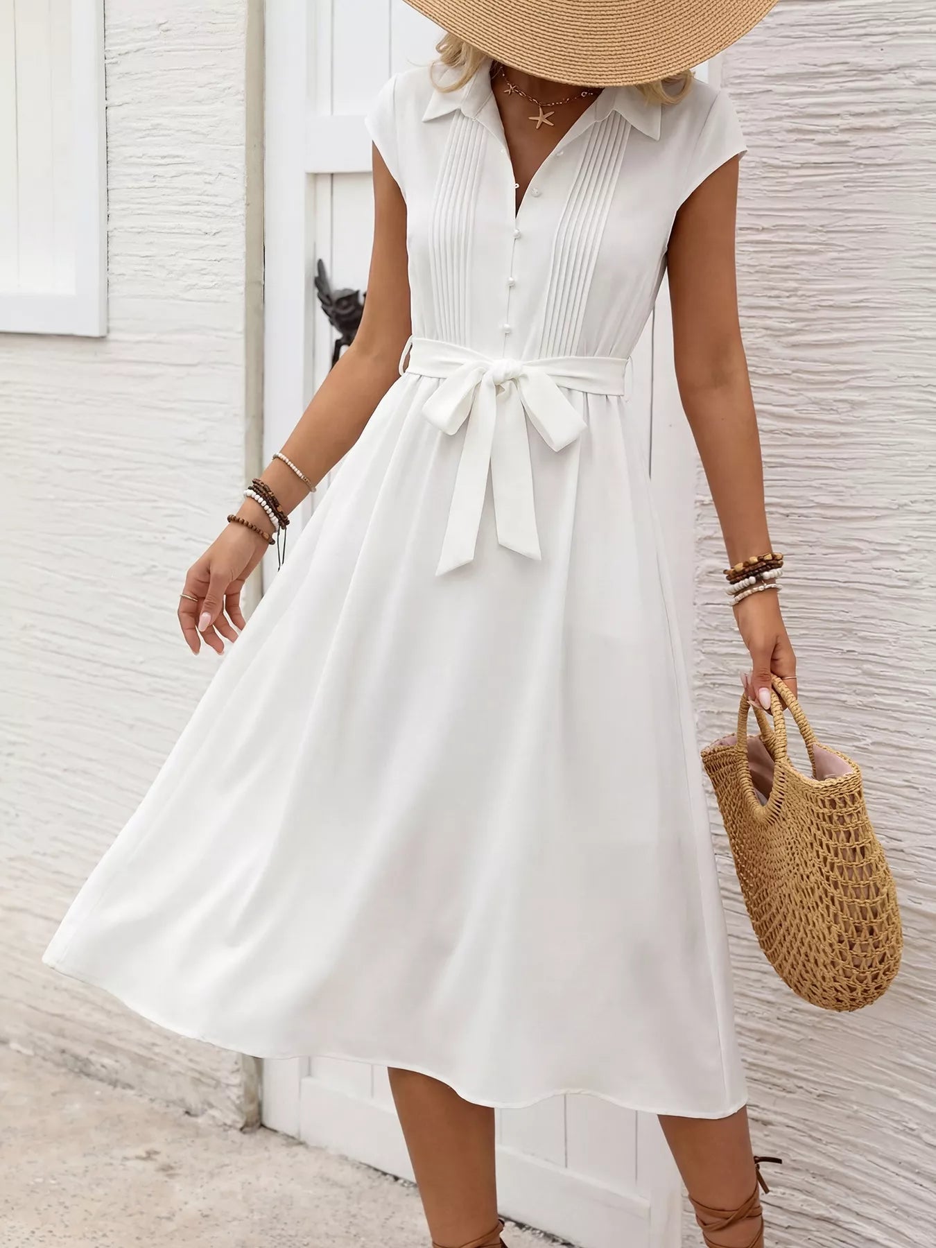 Woman wearing a white dress with a belt, holding a straw bag, against a light background.