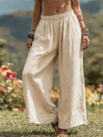 A person outdoors wearing loose, wide-leg linen pants in a light cream or off-white color, standing on grass with blurred flowers and hills in the background.