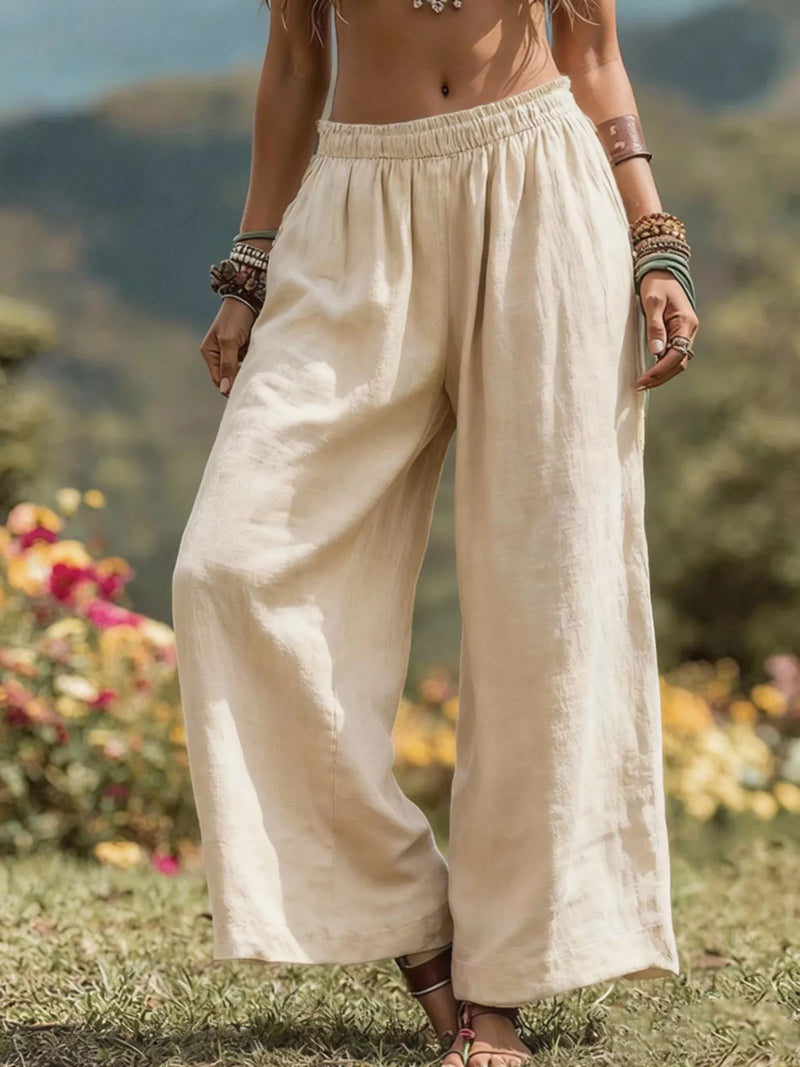 A person outdoors wearing loose, wide-leg linen pants in a light cream or off-white color, standing on grass with blurred flowers and hills in the background.