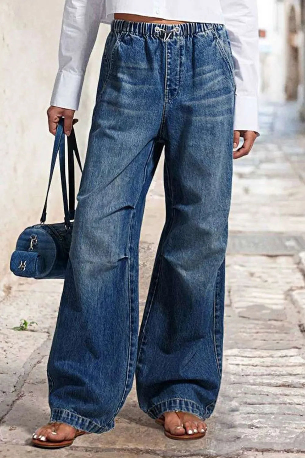 Person wearing wide-leg jeans holding a blue handbag on a stone pavement.