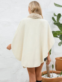 Person wearing a cream-colored poncho with fur trim, standing against a white wall with plants.