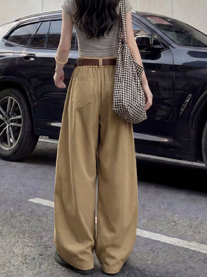 A person standing outdoors in front of a black SUV, wearing loose tan wide-leg pants with a brown belt, a gray sleeveless top, and carrying a black-and-white checkered shoulder bag.
