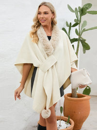 Woman wearing a cream-colored poncho with fur trim, standing next to a potted plant indoors.