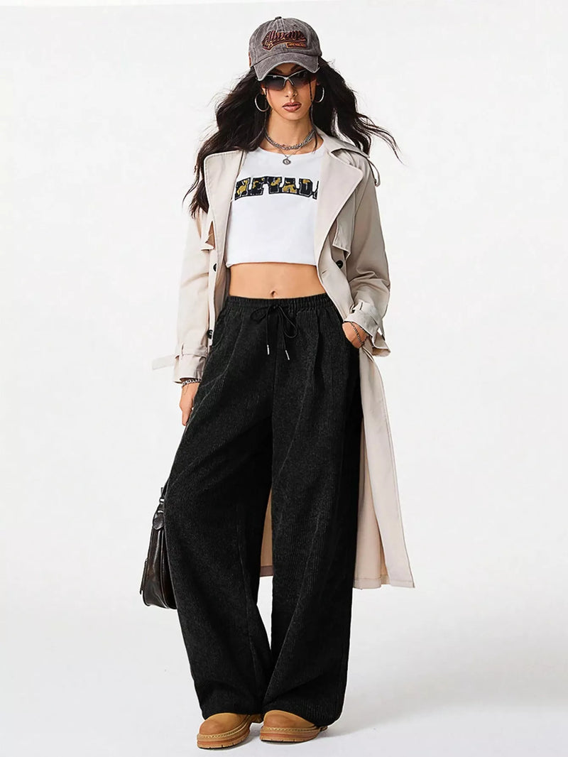 Person wearing a white crop top, black wide-leg pants, beige trench coat, and brown boots on a white background