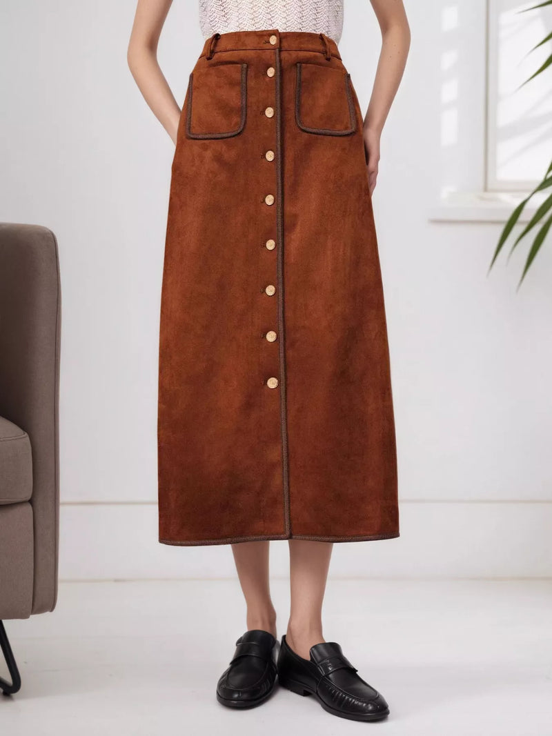 A model wearing a brown suede midi skirt with a button-down front and contrast black leather trim, paired with black loafers in a bright indoor setting.