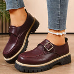Brown leather shoes with buckles worn with blue jeans on a wooden floor.