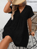 Person wearing a black textured dress with a straw hat against a white brick wall.