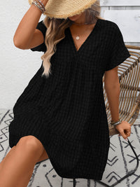 Person wearing a black textured dress with a straw hat against a white brick wall.