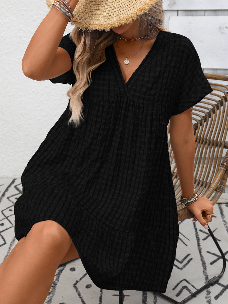 Person wearing a black textured dress with a straw hat against a white brick wall.