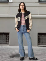 A woman stands outdoors in front of a gray building, wearing a varsity-style jacket with black and beige panels, a striped pink and white top, light blue distressed flare jeans, and black boots.